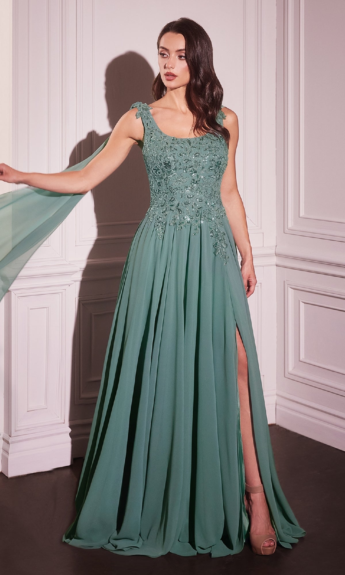 A woman stands in a white-paneled room wearing the Shoulder Sash Long Formal Dress: Ladivine CDS509—an elegant green A-line gown featuring floral embroidery and a high slit.